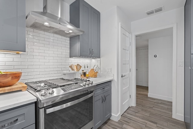 kitchen with gray cabinets, decorative backsplash, gas stove, wall chimney range hood, and light wood-type flooring