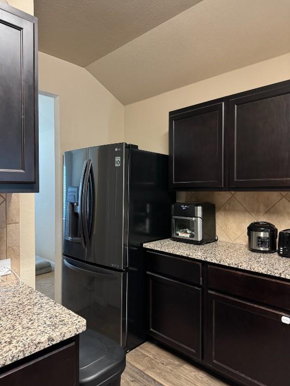 kitchen with lofted ceiling, backsplash, light stone counters, black fridge, and light wood-type flooring