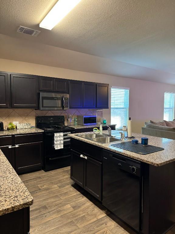 kitchen featuring sink, black appliances, light hardwood / wood-style flooring, a kitchen island with sink, and backsplash