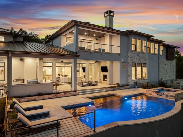 back house at dusk featuring a patio, a balcony, exterior bar, a swimming pool with hot tub, and exterior kitchen