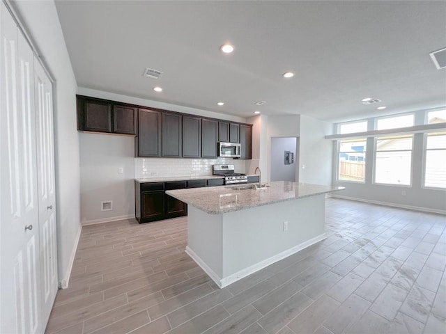 kitchen featuring appliances with stainless steel finishes, an island with sink, sink, backsplash, and light stone countertops