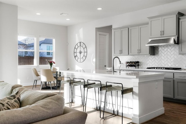 kitchen with light countertops, open floor plan, a kitchen island with sink, wall chimney range hood, and gas cooktop