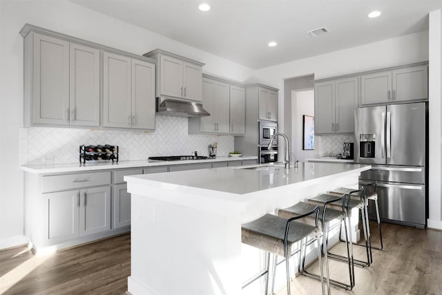 kitchen with a kitchen island with sink, under cabinet range hood, a breakfast bar, light countertops, and appliances with stainless steel finishes