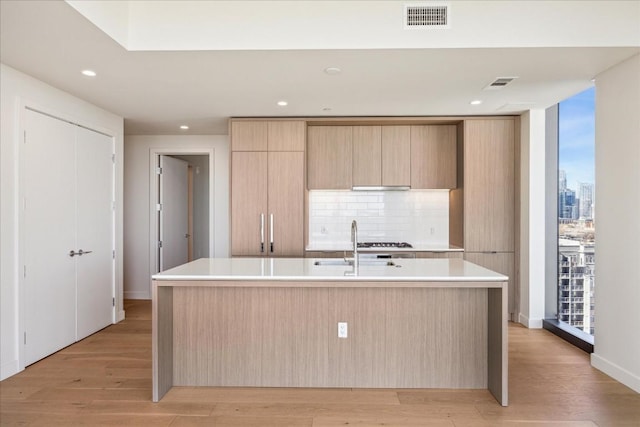 kitchen with light brown cabinetry, sink, an island with sink, light hardwood / wood-style floors, and backsplash