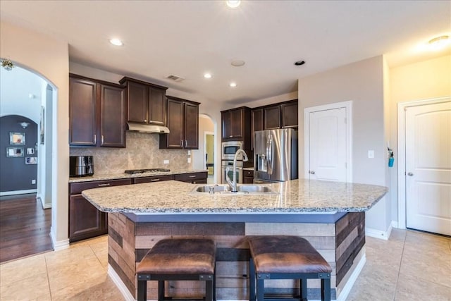 kitchen featuring dark brown cabinetry, a breakfast bar area, appliances with stainless steel finishes, an island with sink, and backsplash