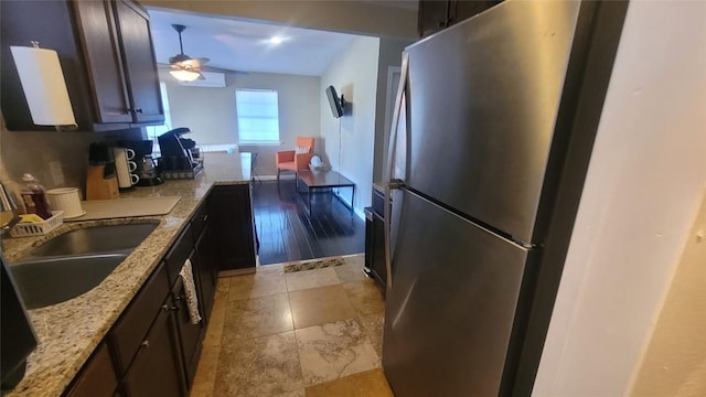 kitchen featuring sink, stainless steel refrigerator, ceiling fan, dark brown cabinets, and light stone countertops