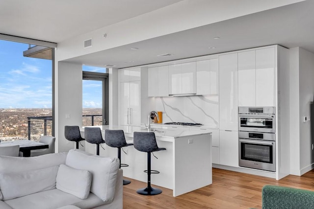 kitchen featuring tasteful backsplash, white cabinets, stainless steel double oven, light hardwood / wood-style floors, and a center island with sink