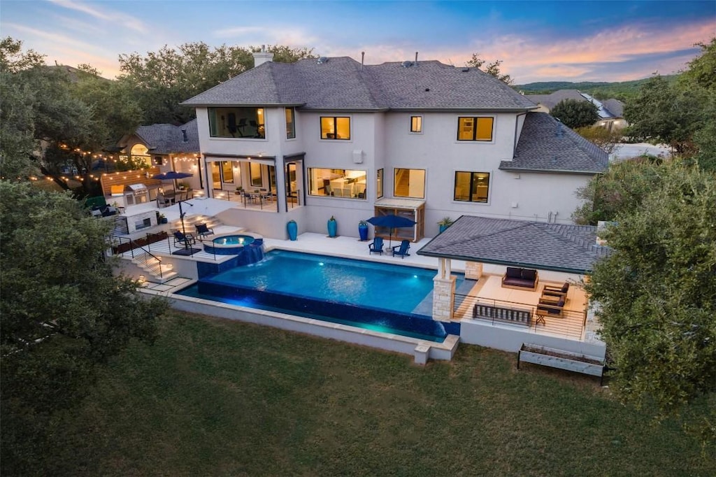 back house at dusk with a lawn, area for grilling, an outdoor living space with a fire pit, a pool with hot tub, and a patio area