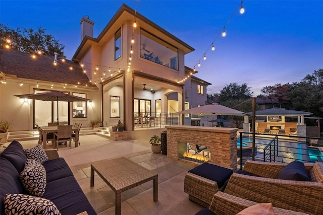 back house at dusk with exterior bar, ceiling fan, an outdoor living space with a fireplace, and a patio area