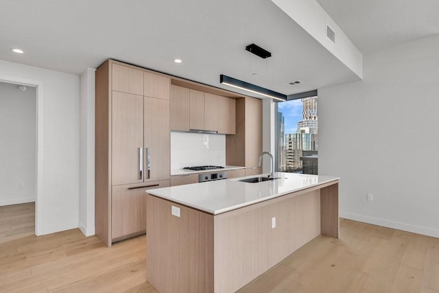 kitchen with sink, stainless steel gas cooktop, a kitchen island with sink, light brown cabinets, and light hardwood / wood-style flooring