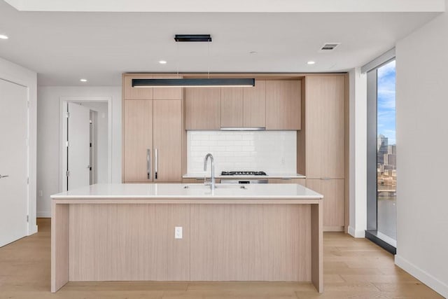 kitchen with light brown cabinetry, hanging light fixtures, a center island with sink, light hardwood / wood-style floors, and backsplash