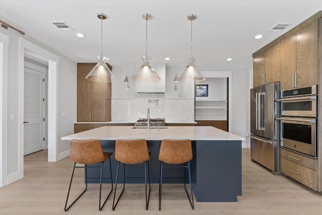 kitchen with appliances with stainless steel finishes, decorative light fixtures, a kitchen bar, a kitchen island with sink, and light wood-type flooring