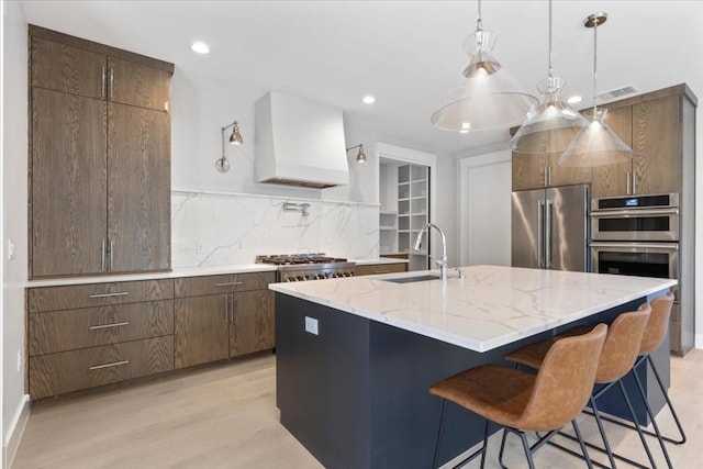 kitchen with premium range hood, sink, hanging light fixtures, stainless steel appliances, and a center island with sink