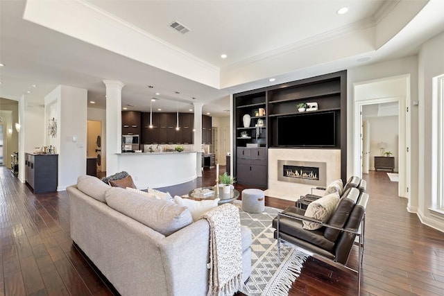 living room featuring built in shelves, dark wood-type flooring, ornate columns, a raised ceiling, and a premium fireplace