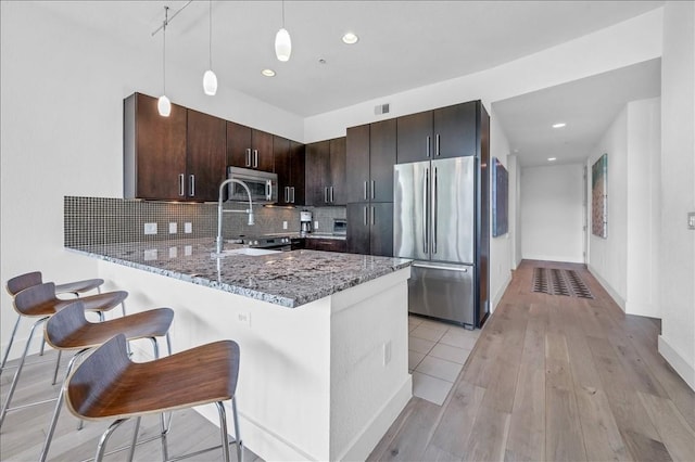 kitchen with dark brown cabinets, hanging light fixtures, kitchen peninsula, stainless steel appliances, and light stone countertops