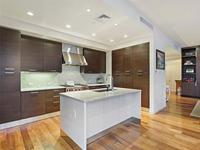 kitchen with sink, a kitchen island with sink, dark brown cabinetry, light wood-type flooring, and wall chimney exhaust hood