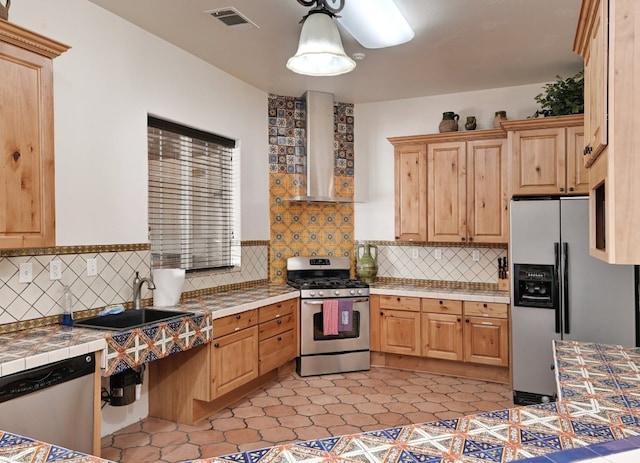 kitchen featuring wall chimney range hood, sink, hanging light fixtures, stainless steel appliances, and tile countertops