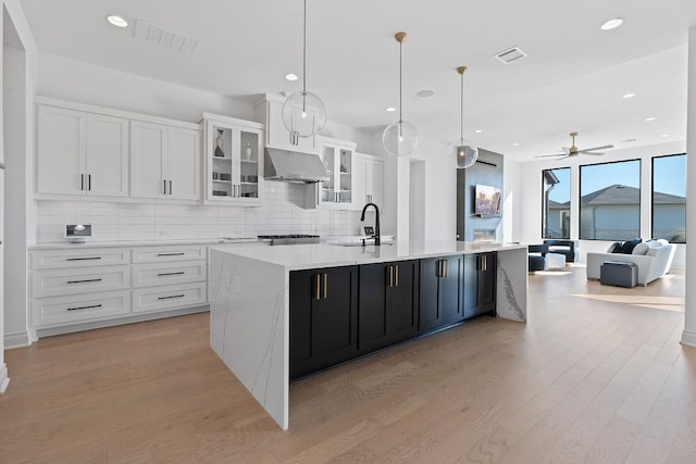 kitchen with white cabinets, a large island with sink, hanging light fixtures, exhaust hood, and light wood-type flooring