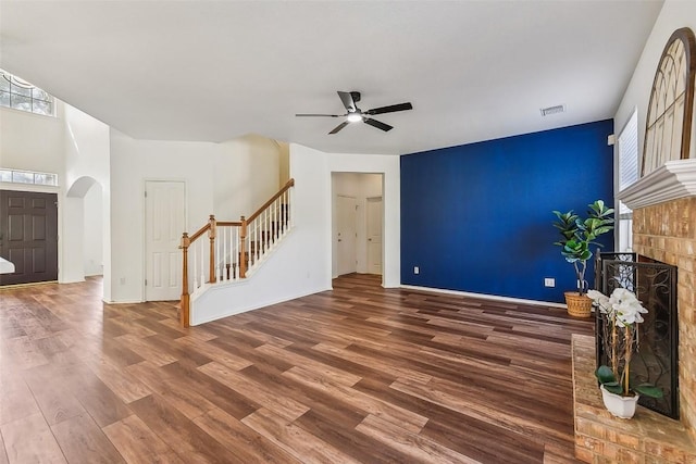 unfurnished living room featuring arched walkways, a brick fireplace, wood finished floors, stairs, and ceiling fan