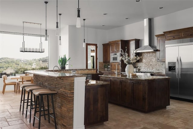 kitchen featuring wall chimney range hood, hanging light fixtures, stainless steel appliances, an island with sink, and dark stone counters