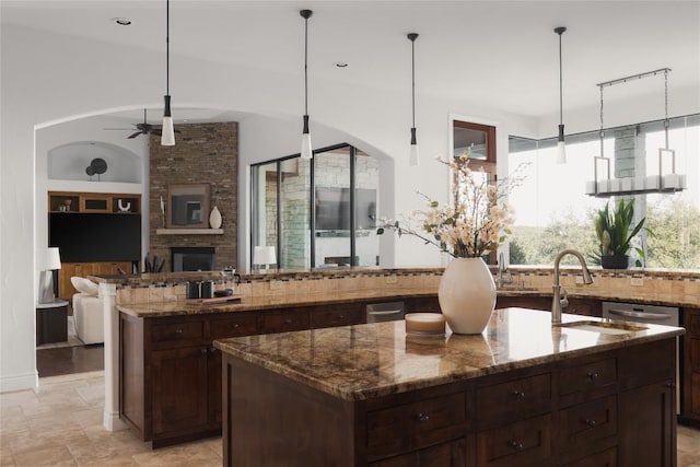 kitchen featuring a stone fireplace, sink, an island with sink, stone counters, and ceiling fan