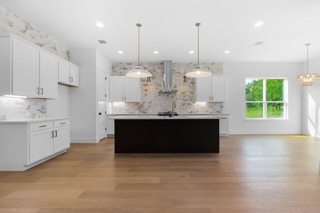 kitchen with white cabinetry, wall chimney range hood, hanging light fixtures, and a center island with sink