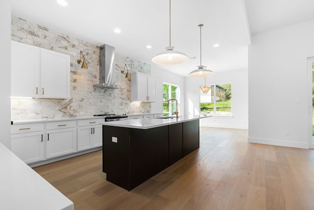 kitchen featuring wall chimney exhaust hood, decorative light fixtures, a center island with sink, and white cabinets