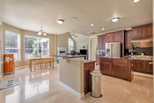 kitchen with ceiling fan with notable chandelier, an island with sink, dark stone countertops, exhaust hood, and stainless steel refrigerator with ice dispenser