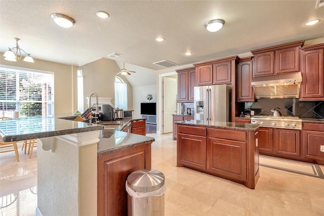 kitchen with appliances with stainless steel finishes, ceiling fan with notable chandelier, dark stone counters, decorative backsplash, and a center island with sink