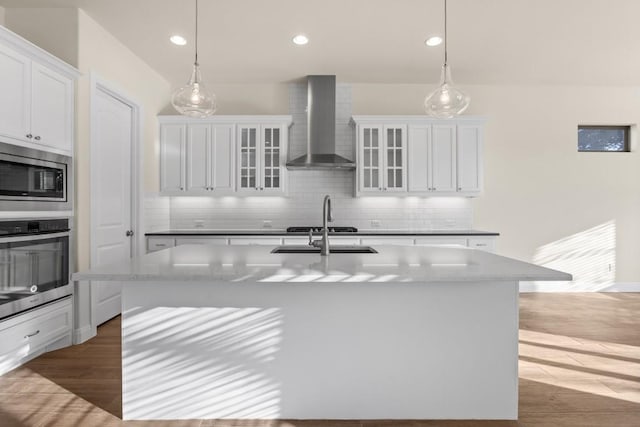 kitchen featuring wall chimney exhaust hood, decorative light fixtures, appliances with stainless steel finishes, an island with sink, and white cabinets