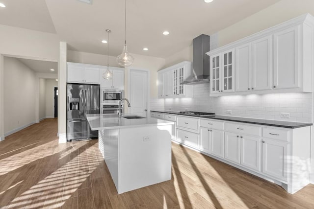 kitchen featuring hanging light fixtures, stainless steel appliances, an island with sink, white cabinets, and wall chimney exhaust hood