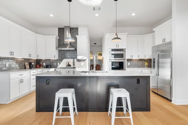 kitchen featuring an island with sink, white cabinetry, a kitchen breakfast bar, stainless steel appliances, and wall chimney range hood