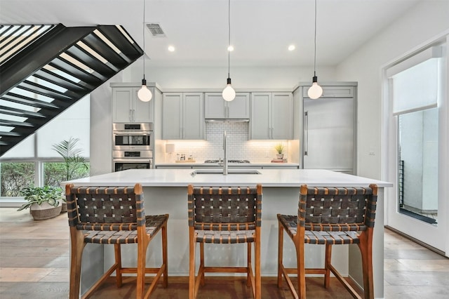 kitchen featuring stainless steel appliances, decorative light fixtures, gray cabinets, and a center island with sink