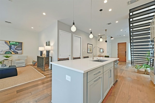 kitchen with pendant lighting, dishwasher, an island with sink, sink, and light hardwood / wood-style floors