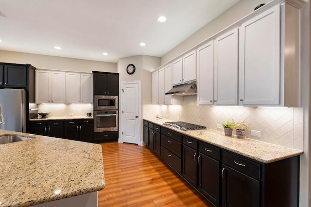 kitchen with appliances with stainless steel finishes, white cabinetry, backsplash, light stone countertops, and light wood-type flooring