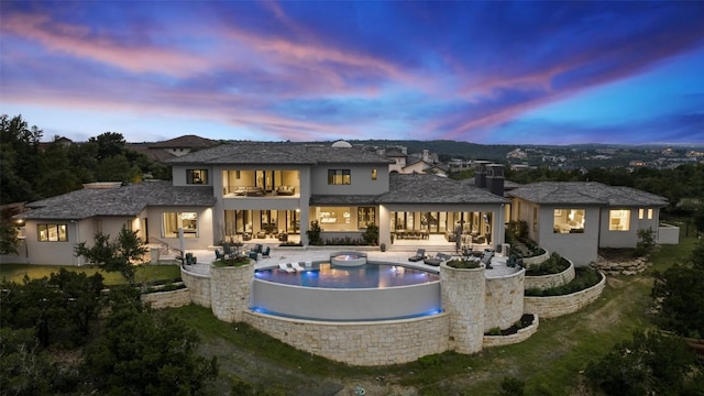 back of property at dusk featuring stucco siding, an infinity pool, a patio, an outdoor hangout area, and a balcony