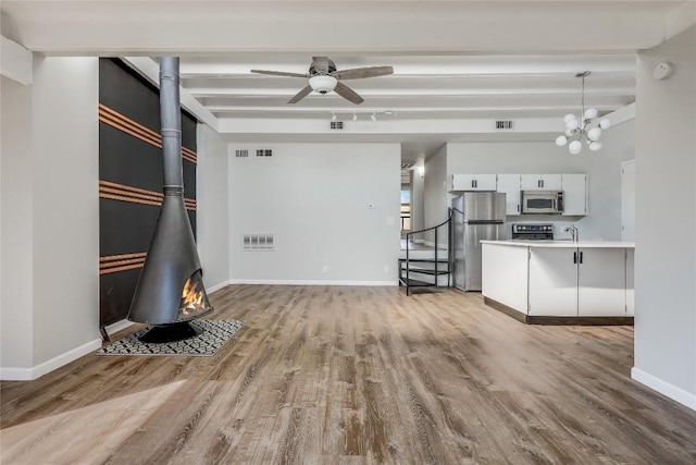 unfurnished living room with beamed ceiling, a wood stove, ceiling fan with notable chandelier, and light hardwood / wood-style flooring