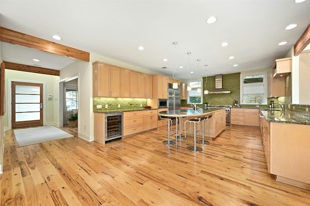 kitchen featuring light brown cabinetry, an island with sink, sink, wine cooler, and wall chimney range hood