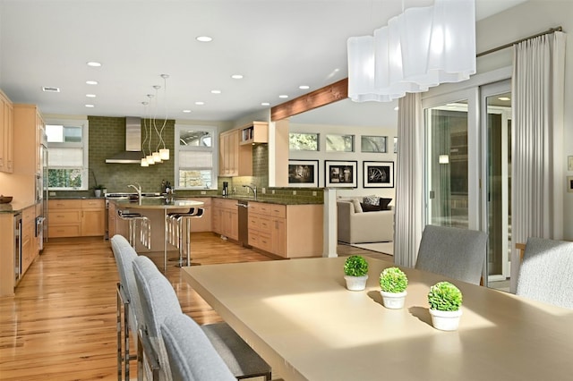 kitchen featuring wall chimney exhaust hood, tasteful backsplash, light brown cabinets, a kitchen breakfast bar, and light hardwood / wood-style floors