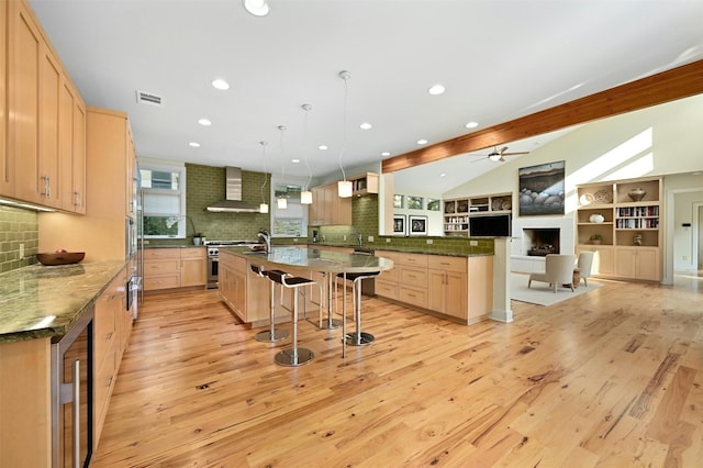 kitchen with pendant lighting, vaulted ceiling with beams, stainless steel range, light brown cabinetry, and wall chimney exhaust hood