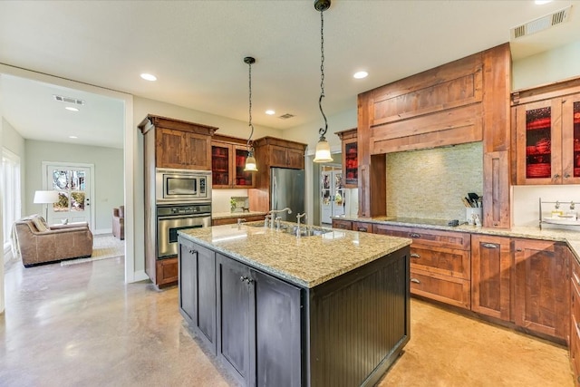 kitchen with light stone counters, decorative light fixtures, appliances with stainless steel finishes, a kitchen island with sink, and backsplash