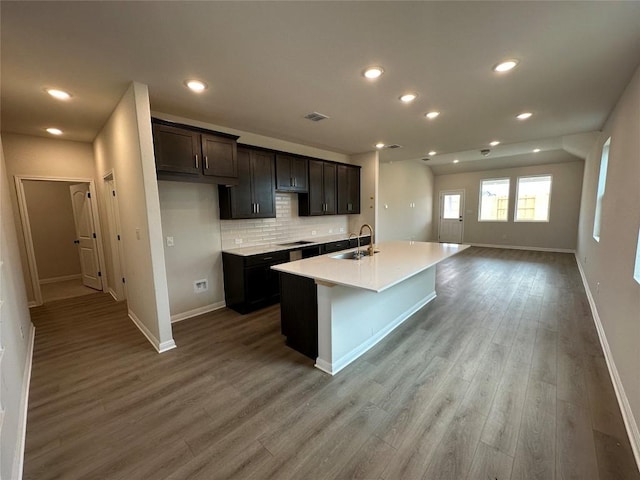 kitchen with an island with sink, a sink, backsplash, wood finished floors, and baseboards