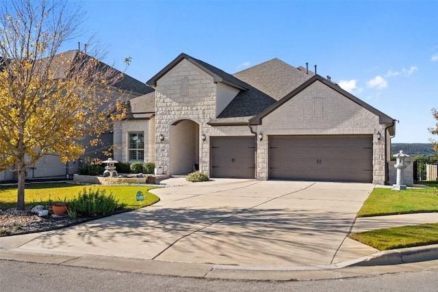 french country style house featuring concrete driveway, stone siding, roof with shingles, an attached garage, and a front lawn