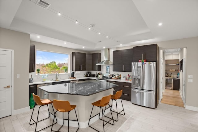 kitchen with wall chimney range hood, a breakfast bar area, appliances with stainless steel finishes, a center island, and a tray ceiling