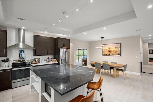 kitchen with a kitchen island, wall chimney range hood, appliances with stainless steel finishes, light wood-type flooring, and a tray ceiling