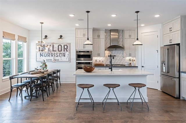 kitchen featuring stainless steel appliances, a kitchen island with sink, wall chimney range hood, and decorative light fixtures