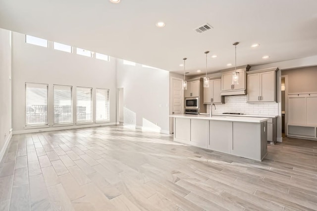 kitchen with hanging light fixtures, a center island with sink, stainless steel microwave, gray cabinets, and a wealth of natural light