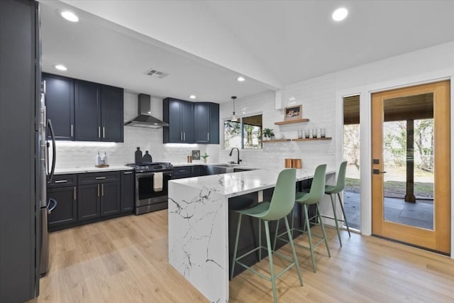 kitchen featuring a kitchen bar, light stone counters, kitchen peninsula, stainless steel appliances, and wall chimney range hood