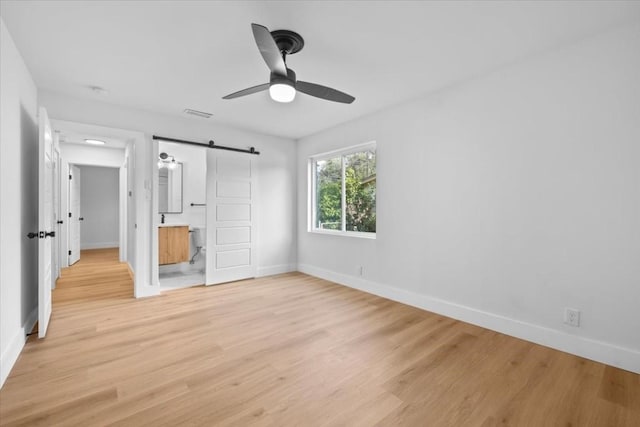 unfurnished bedroom featuring ceiling fan, ensuite bathroom, a barn door, and light wood-type flooring