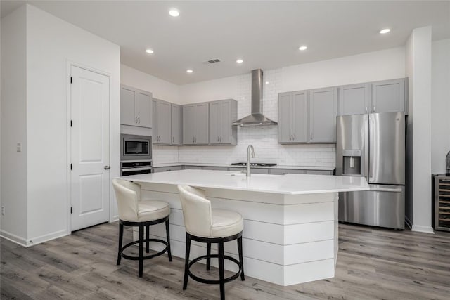 kitchen with wall chimney exhaust hood, appliances with stainless steel finishes, a kitchen island with sink, and light hardwood / wood-style flooring